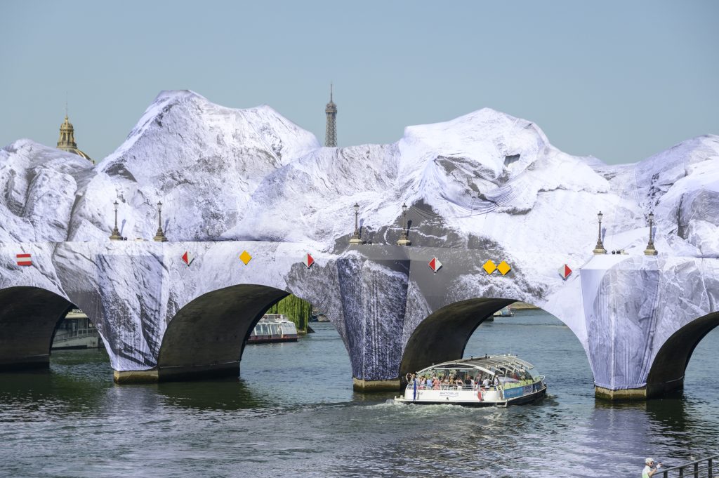 Photo de rue capturant de face deux arches du Pont-Neuf à Paris, métamorphosé par l'installation de JR, « La Caverne du Pont Neuf ». La bâche en trompe-l'œil donne au monument historique l'apparence d'un massif montagneux escarpé s'élevant au-dessus de l'eau. Une grande péniche de fret commerciale bleue et blanche navigue sur la Seine et s'apprête à passer sous l'une des arches du pont. Le ciel en arrière-plan est totalement épuré et d'un bleu très clair.