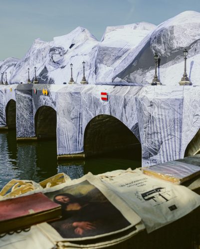 Photo de rue à Paris montrant une vue en contre-plongée et de trois-quarts du Pont-Neuf, entièrement recouvert par une installation artistique monumentale de JR intitulée « La Caverne du Pont Neuf ». Le pont est drapé d'une immense bâche imprimée en trompe-l'œil représentant une montagne rocheuse en noir et blanc, dont les reliefs épousent les arches. Au premier plan, légèrement flou, se trouve le stand d'un bouquiniste des quais de Seine, où sont exposés des sacs en toile (tote bags), dont l'un arbore une reproduction de la Joconde. L'eau de la Seine reflète la structure du pont sous un ciel bleu et dégagé.