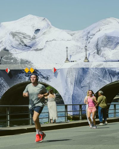 Scène de vie et photo de rue animée sur les quais de Seine à Paris. Au premier plan, un homme en short sombre et t-shirt gris court vers la gauche, le mouvement créant un léger flou. En arrière-plan, d'autres piétons et joggeurs longent la rambarde. Derrière eux se dresse le Pont-Neuf, entièrement enveloppé par l'œuvre monumentale de JR, « La Caverne du Pont Neuf », simulant un paysage de roche et de glace. Les lampadaires du pont émergent de cette structure textile blanche et grise.