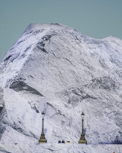 Gros plan vertical et graphique sur une section de l'installation artistique de JR, « La Caverne du Pont Neuf », à Paris. La bâche imprimée s'élève très haut dans le cadre, imitant parfaitement le sommet pointu et les parois abruptes d'une montagne enneigée. Seuls deux lampadaires parisiens traditionnels en fonte, émergeant à la base de la structure, trahissent la présence du pont sous la bâche. Le ciel en arrière-plan est d'un bleu uni et pâle.