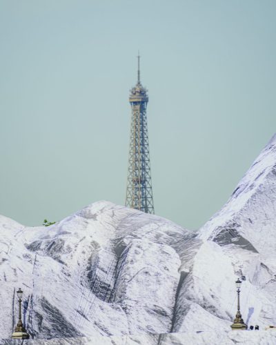 Composition photographique superposant deux symboles parisiens. Au premier plan, les faux sommets montagneux en trompe-l'œil de l'installation de JR, « La Caverne du Pont Neuf », recouvrent le pont. Juste derrière ces reliefs de toile blanche et grise, la partie supérieure de la Tour Eiffel se dresse verticalement dans un ciel clair, créant un contraste insolite entre l'architecture de fer et le paysage alpin artificiel.
