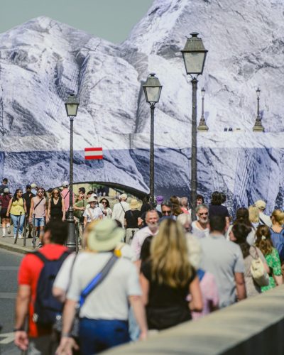 Photo de rue prise en longue focale montrant une foule dense de dos et de face marchant sur un quai à Paris. En arrière-plan, l'immense bâche de l'œuvre de JR, « La Caverne du Pont Neuf », habille les structures du Pont-Neuf, créant un décor de montagne qui surplombe les passants. Plusieurs lampadaires parisiens s'alignent le long du garde-corps au milieu de la foule.