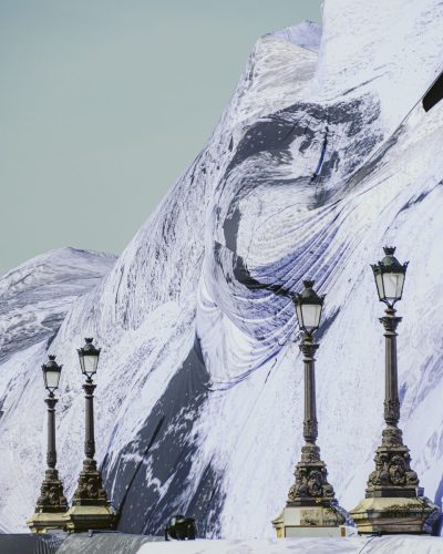 Prise de vue en contre-plongée mettant l'accent sur les détails de l'œuvre de JR, « La Caverne du Pont Neuf », à Paris. La bâche drapée révèle des textures complexes de roches, de crevasses et de neige en noir et blanc. Quatre lampadaires historiques alignés au premier plan semblent grimper le long de la paroi de cette montagne artificielle. Le ciel lumineux occupe le coin supérieur gauche.