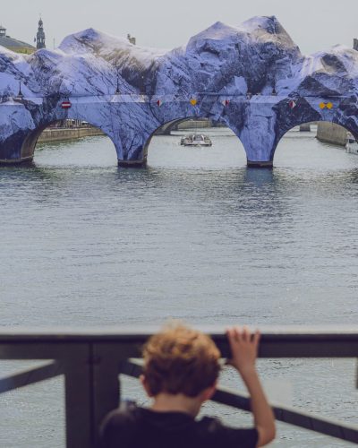 Depuis le Pont des Arts, un jeune enfant vu de dos et flouté au premier plan s'appuie contre une rambarde métallique, regardant le fleuve. Au centre de l'image, un bateau-mouche rempli de touristes navigue sur l'eau en direction du Pont-Neuf. Le pont est entièrement recouvert par la bâche de JR, « La Caverne du Pont Neuf », transformant le paysage urbain de Paris en une vallée fluviale au pied de falaises rocheuses.