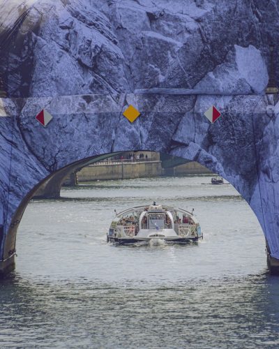 Plan serré et centré sous l'arche du Pont-Neuf à Paris, modifié par l'installation « La Caverne du Pont Neuf » de JR. La bâche imprimée façon roche entoure l'arche comme l'entrée d'une véritable grotte en pierre. À travers cette ouverture, on voit un bateau de croisière fluviale (« Dauphine ») blanc approcher sur la Seine, avec des passagers visibles sur le pont supérieur.