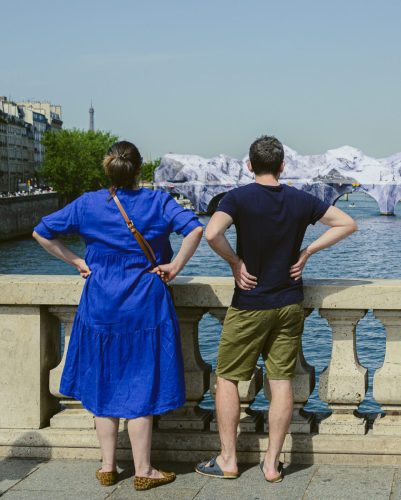 Photo de rue à Paris montrant un couple de dos, les mains sur les hanches, observant le paysage depuis un pont. À gauche, une femme porte une robe bleu vif et des chaussures léopard ; à droite, un homme porte un t-shirt bleu marine et un short kaki. Ils contemplent le Pont-Neuf, situé plus loin sur la Seine, transformé par la bâche de JR en montagne artificielle (« La Caverne du Pont Neuf »). Au loin, à gauche, la silhouette de la Tour Eiffel se dessine discrètement à l'horizon.